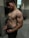 A shirtless man with muscular build standing in a gym locker room. The lighting highlights his physique, and he is wearing dark shorts with visible brand underwear.