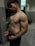 A shirtless man with muscular build standing in a gym locker room. The lighting highlights his physique, and he is wearing dark shorts with visible brand underwear.