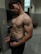 A shirtless man with muscular build standing in a gym locker room. The lighting highlights his physique, and he is wearing dark shorts with visible brand underwear.