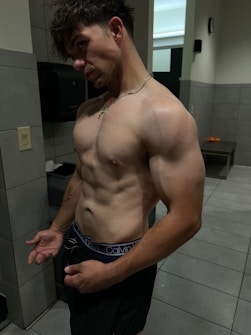 A Shirtless Man With Muscular Build Standing In A Gym Locker Room. The Lighting Highlights His Physique, And He Is Wearing Dark Shorts With Visible Brand Underwear.