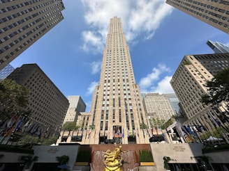 A tall, iconic skyscraper stands prominently in the center, flanked by other high-rise buildings under a clear blue sky with scattered white clouds. At the bottom, a bronze statue and a row of various national flags add to the lively atmosphere.