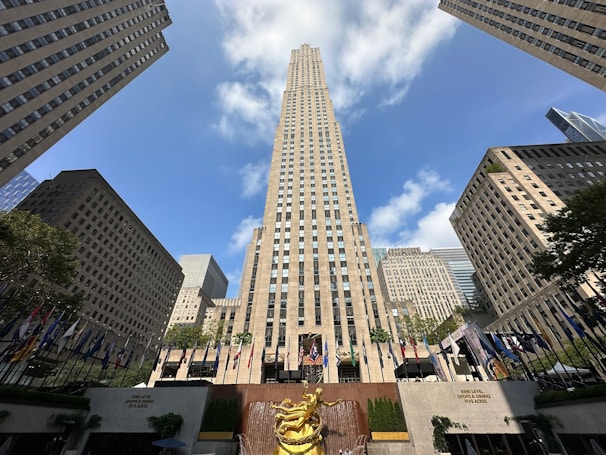 A tall, iconic skyscraper stands prominently in the center, flanked by other high-rise buildings under a clear blue sky with scattered white clouds. At the bottom, a bronze statue and a row of various national flags add to the lively atmosphere.
