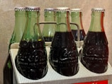 A pack of six green glass bottles filled with a dark liquid, likely a carbonated beverage, secured in a cardboard carrier. The bottles have metal caps and embossed branding on the glass. The lighting highlights the texture of the condensation on the bottles. The background is a plain neutral tone.
