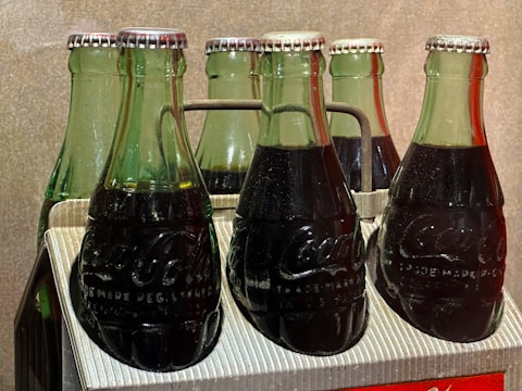 A pack of six green glass bottles filled with a dark liquid, likely a carbonated beverage, secured in a cardboard carrier. The bottles have metal caps and embossed branding on the glass. The lighting highlights the texture of the condensation on the bottles. The background is a plain neutral tone.