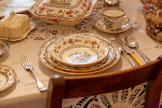 An elegant table setting featuring intricately decorated porcelain plates, bowls, and cups with floral patterns placed on a lace tablecloth. The dining setup includes silverware consisting of a fork, knife, and spoon with handles of varying colors. A serving dish with a lid and a sugar bowl are part of the arrangement.