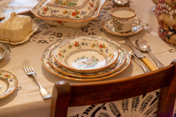 An elegant table setting featuring intricately decorated porcelain plates, bowls, and cups with floral patterns placed on a lace tablecloth. The dining setup includes silverware consisting of a fork, knife, and spoon with handles of varying colors. A serving dish with a lid and a sugar bowl are part of the arrangement.