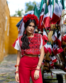 A woman dressed in vibrant traditional clothing stands with a backdrop of multiple Mexican flags. She wears red pants and a decorative top with red, white, and black patterns. Her hair is adorned with red flowers, and she accessorizes with red earrings and bracelets. The scene has a colorful and festive atmosphere.