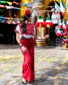 A woman wearing vibrant red and white traditional clothing, adorned with floral accessories, stands amidst festive decorations. Brightly colored paper banners and flags are hung around, creating a lively atmosphere. The setting appears to be an outdoor courtyard, with a yellow and white building in the background.