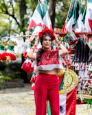 A woman wearing a traditional embroidered blouse and red pants stands among a variety of Mexican-themed decorations and flags. She adjusts a floral headdress on her head, surrounded by vibrant colors and patterns.