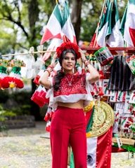 A woman wearing a traditional embroidered blouse and red pants stands among a variety of Mexican-themed decorations and flags. She adjusts a floral headdress on her head, surrounded by vibrant colors and patterns.