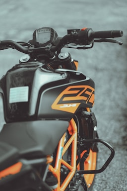 A close-up view of a motorcycle with a focus on the dashboard, handlebars, and part of the frame. The body features black and orange colors, with the number 250 prominently displayed. The motorcycle has a sporty design and is parked on a gray, textured surface.