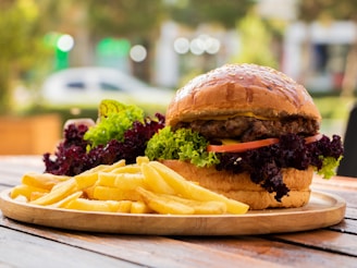 A large hamburger filled with lettuce, tomatoes, and a beef patty is placed on a wooden plate next to a serving of golden French fries. The setting appears outdoors with a blurred background of greenery and potential urban elements, suggesting a casual dining atmosphere.
