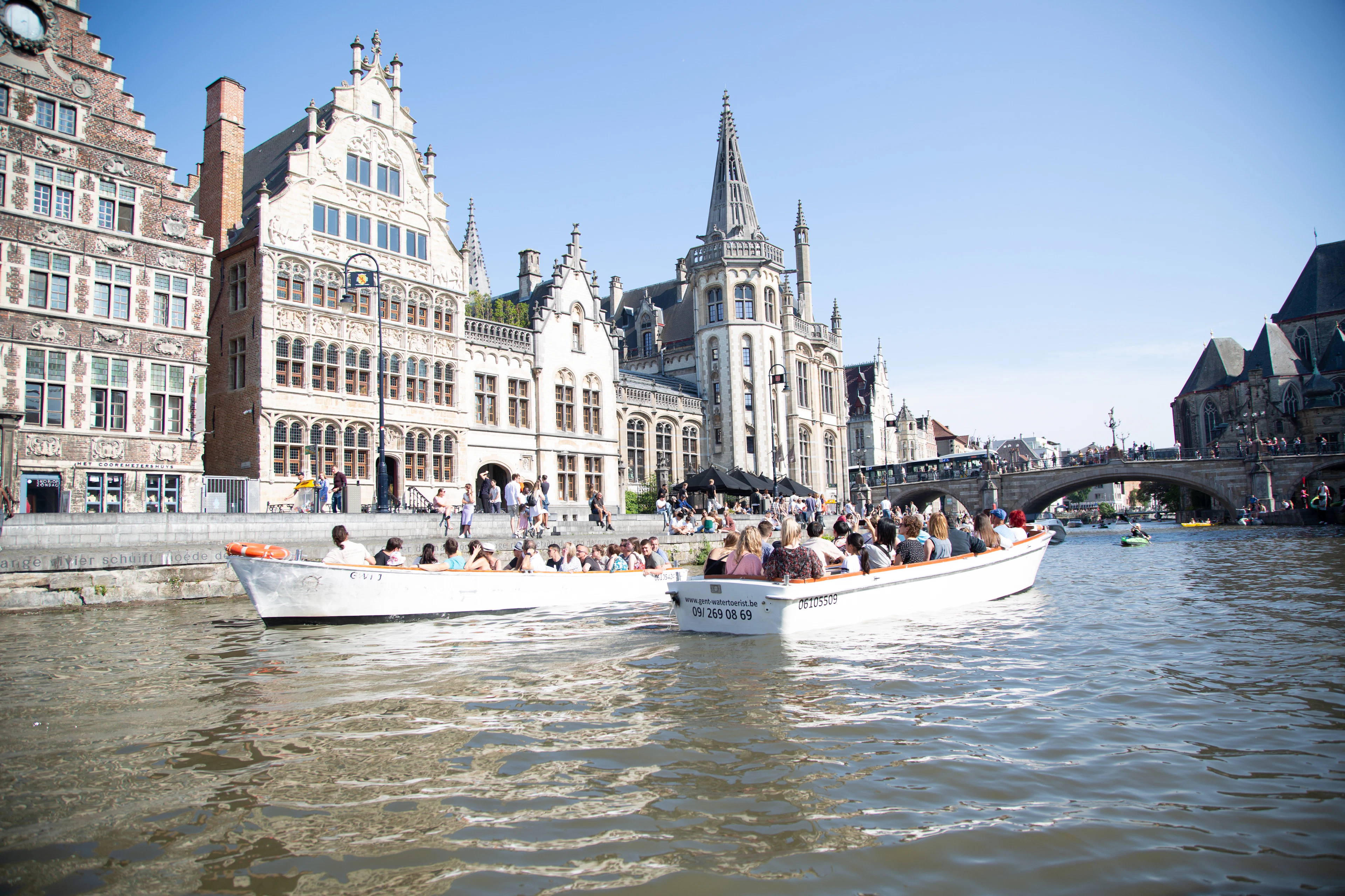 Two open tourist boats full of passengers on Ghent's canal in front of the stepped-gable guild houses of the Graslei, with a stone bridge downstream