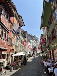 A cobblestone street lined with colorful buildings displaying multiple Swiss flags. Outdoor diners are seated at tables along the street, enjoying a sunny day. People walk leisurely, creating a lively yet relaxed atmosphere.