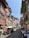 A cobblestone street lined with colorful buildings displaying multiple Swiss flags. Outdoor diners are seated at tables along the street, enjoying a sunny day. People walk leisurely, creating a lively yet relaxed atmosphere.