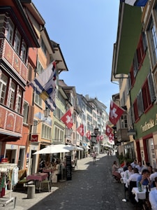 A cobblestone street lined with colorful buildings displaying multiple Swiss flags. Outdoor diners are seated at tables along the street, enjoying a sunny day. People walk leisurely, creating a lively yet relaxed atmosphere.