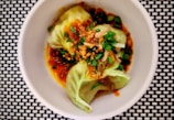 A neatly presented dish featuring dumplings garnished with chopped green onions and crispy fried shallots. The dumplings are nestled in a rich, reddish-brown sauce and are placed in a white bowl, which contrasts with the black and white checkered surface below.