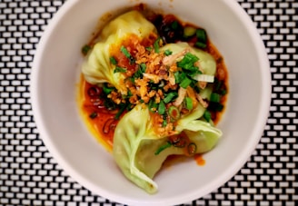 A neatly presented dish featuring dumplings garnished with chopped green onions and crispy fried shallots. The dumplings are nestled in a rich, reddish-brown sauce and are placed in a white bowl, which contrasts with the black and white checkered surface below.