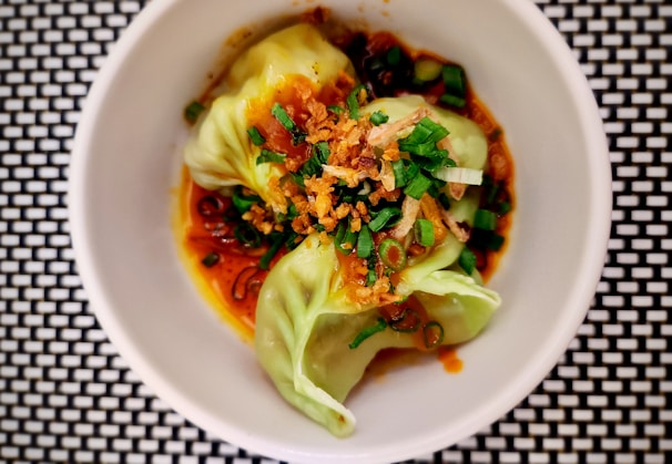A neatly presented dish featuring dumplings garnished with chopped green onions and crispy fried shallots. The dumplings are nestled in a rich, reddish-brown sauce and are placed in a white bowl, which contrasts with the black and white checkered surface below.