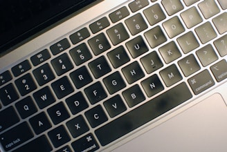 A close-up view of a laptop keyboard with black keys and white letters. The keyboard features shortcut icons and a matte finish.