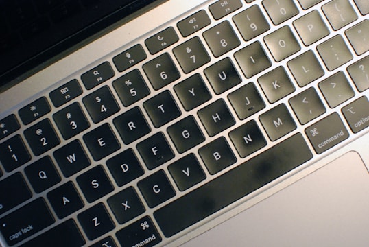 A close-up view of a laptop keyboard with black keys and white letters. The keyboard features shortcut icons and a matte finish.