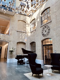 A stylish interior space with high ceilings and large windows, featuring a brick wall. A grand piano is positioned in the corner next to a modern sculpture-like lighting fixture hanging from the ceiling. Two tall, black armchairs and a small white table are arranged on a rug in the foreground. A large decorative wall clock adds to the sophisticated ambiance.