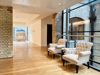 A modern interior space featuring two elegant, striped armchairs positioned on a polished wooden floor. The area is illuminated by natural light coming through large glass windows that reveal an exposed brick wall exterior. A hallway extends into another section, showcasing a cushioned bench underneath an arched window.