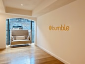 A modern, minimalist office space with a cozy beige sofa placed near large windows featuring a view of a brick building facade. The sofa has decorative pillows with colorful patterns. The wall is adorned with the word 'bumble' in bold yellow letters, accompanied by a hexagonal logo. Wooden flooring adds warmth to the otherwise neutral color palette of the room.
