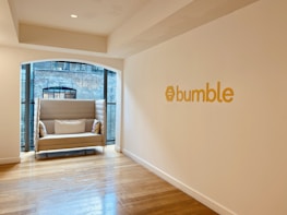 A modern, minimalist office space with a cozy beige sofa placed near large windows featuring a view of a brick building facade. The sofa has decorative pillows with colorful patterns. The wall is adorned with the word 'bumble' in bold yellow letters, accompanied by a hexagonal logo. Wooden flooring adds warmth to the otherwise neutral color palette of the room.