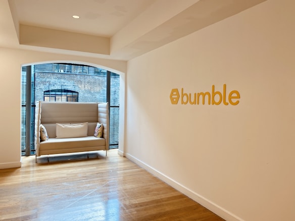 A modern, minimalist office space with a cozy beige sofa placed near large windows featuring a view of a brick building facade. The sofa has decorative pillows with colorful patterns. The wall is adorned with the word 'bumble' in bold yellow letters, accompanied by a hexagonal logo. Wooden flooring adds warmth to the otherwise neutral color palette of the room.
