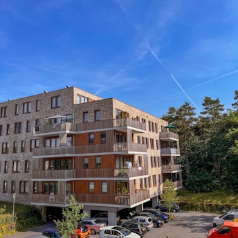 A modern apartment building with a combination of brick and wooden paneling, featuring balconies with railings. The building is surrounded by a parking area with several cars and is set against a backdrop of trees and a clear blue sky.