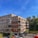 A modern apartment building with a combination of brick and wooden paneling, featuring balconies with railings. The building is surrounded by a parking area with several cars and is set against a backdrop of trees and a clear blue sky.