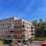 A modern apartment building with a combination of brick and wooden paneling, featuring balconies with railings. The building is surrounded by a parking area with several cars and is set against a backdrop of trees and a clear blue sky.