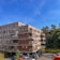 A modern apartment building with a combination of brick and wooden paneling, featuring balconies with railings. The building is surrounded by a parking area with several cars and is set against a backdrop of trees and a clear blue sky.