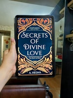 A hand is holding an electronic device displaying a book cover titled 'Secrets of Divine Love' by A. Helwa. The cover features ornate, colorful patterns with floral designs in shades of gold, orange, and red on a dark blue background. The setting appears to be indoors, with a modern, comfortable living area in the background.