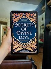 A hand is holding an electronic device displaying a book cover titled 'Secrets of Divine Love' by A. Helwa. The cover features ornate, colorful patterns with floral designs in shades of gold, orange, and red on a dark blue background. The setting appears to be indoors, with a modern, comfortable living area in the background.