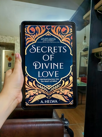A hand is holding an electronic device displaying a book cover titled 'Secrets of Divine Love' by A. Helwa. The cover features ornate, colorful patterns with floral designs in shades of gold, orange, and red on a dark blue background. The setting appears to be indoors, with a modern, comfortable living area in the background.
