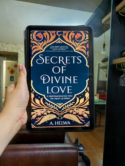 A hand is holding an electronic device displaying a book cover titled 'Secrets of Divine Love' by A. Helwa. The cover features ornate, colorful patterns with floral designs in shades of gold, orange, and red on a dark blue background. The setting appears to be indoors, with a modern, comfortable living area in the background.