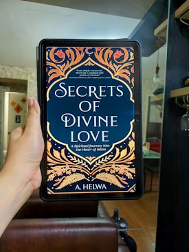 A hand is holding an electronic device displaying a book cover titled 'Secrets of Divine Love' by A. Helwa. The cover features ornate, colorful patterns with floral designs in shades of gold, orange, and red on a dark blue background. The setting appears to be indoors, with a modern, comfortable living area in the background.
