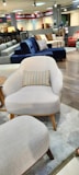 A furniture showroom with various sofas and chairs on display. In the foreground, a light gray armchair with wooden legs and a multicolored throw pillow is prominently visible. Surrounding the armchair are a few other sofas in different colors, including blue and red, set on a wooden floor. The background features shelves with fabric samples and additional seating options.