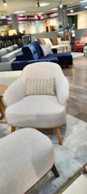 A furniture showroom with various sofas and chairs on display. In the foreground, a light gray armchair with wooden legs and a multicolored throw pillow is prominently visible. Surrounding the armchair are a few other sofas in different colors, including blue and red, set on a wooden floor. The background features shelves with fabric samples and additional seating options.