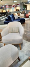 A furniture showroom with various sofas and chairs on display. In the foreground, a light gray armchair with wooden legs and a multicolored throw pillow is prominently visible. Surrounding the armchair are a few other sofas in different colors, including blue and red, set on a wooden floor. The background features shelves with fabric samples and additional seating options.