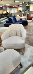 A furniture showroom with various sofas and chairs on display. In the foreground, a light gray armchair with wooden legs and a multicolored throw pillow is prominently visible. Surrounding the armchair are a few other sofas in different colors, including blue and red, set on a wooden floor. The background features shelves with fabric samples and additional seating options.