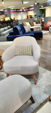 A furniture showroom with various sofas and chairs on display. In the foreground, a light gray armchair with wooden legs and a multicolored throw pillow is prominently visible. Surrounding the armchair are a few other sofas in different colors, including blue and red, set on a wooden floor. The background features shelves with fabric samples and additional seating options.
