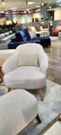 A furniture showroom with various sofas and chairs on display. In the foreground, a light gray armchair with wooden legs and a multicolored throw pillow is prominently visible. Surrounding the armchair are a few other sofas in different colors, including blue and red, set on a wooden floor. The background features shelves with fabric samples and additional seating options.