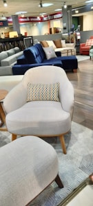 A furniture showroom with various sofas and chairs on display. In the foreground, a light gray armchair with wooden legs and a multicolored throw pillow is prominently visible. Surrounding the armchair are a few other sofas in different colors, including blue and red, set on a wooden floor. The background features shelves with fabric samples and additional seating options.