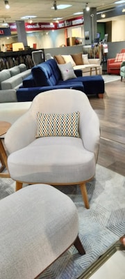 A furniture showroom with various sofas and chairs on display. In the foreground, a light gray armchair with wooden legs and a multicolored throw pillow is prominently visible. Surrounding the armchair are a few other sofas in different colors, including blue and red, set on a wooden floor. The background features shelves with fabric samples and additional seating options.