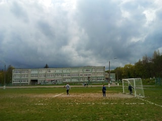 A school building under a cloudy sky with a football field in the foreground. Several people are present on the field, including children playing near a goalpost. The setting appears to be outdoors within a schoolyard environment surrounded by trees.
