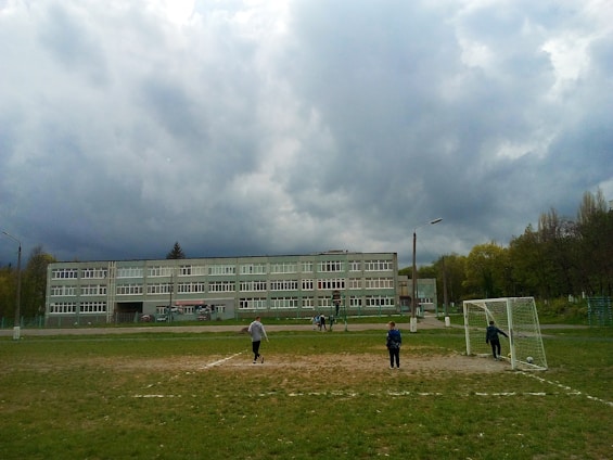 A school building under a cloudy sky with a football field in the foreground. Several people are present on the field, including children playing near a goalpost. The setting appears to be outdoors within a schoolyard environment surrounded by trees.