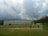 A school building under a cloudy sky with a football field in the foreground. Several people are present on the field, including children playing near a goalpost. The setting appears to be outdoors within a schoolyard environment surrounded by trees.